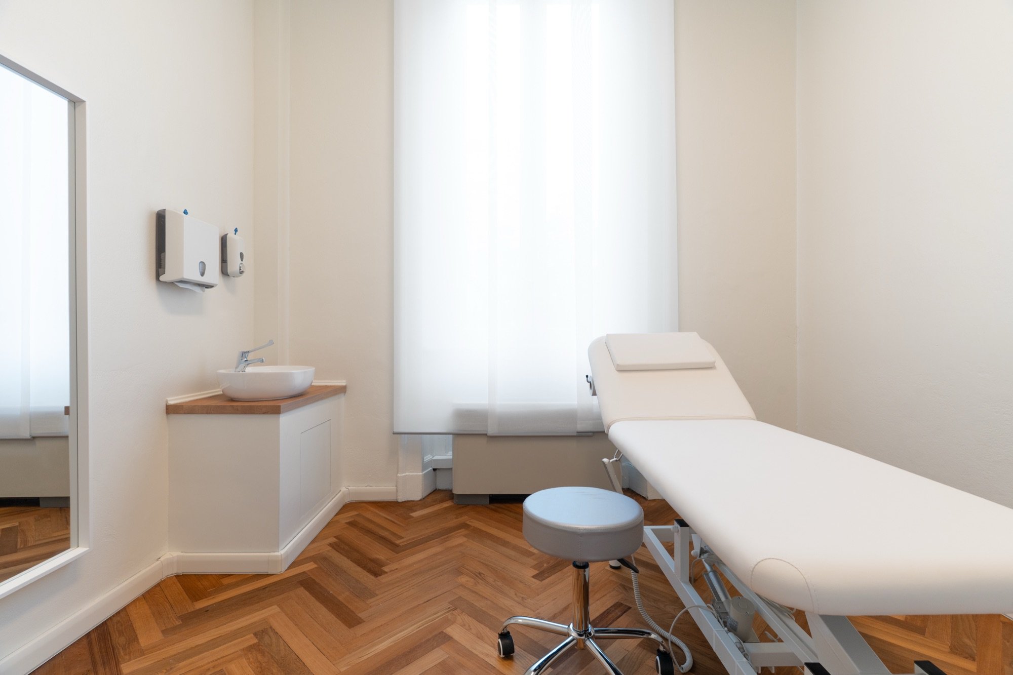 Lettino da visita con sgabelli e lavabo clinico vicino alla finestra in studio professionale Standard presso Spatium Corso Vercelli, Milano